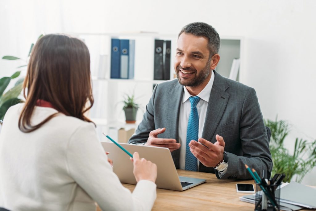 When To Hire A Student Loan Professional A smiling businessman in a grey suit and blue tie gestures while speaking to a client in a bright office. This image represents the role of financial professionals—specifically Certified Financial Planners (CFPs) or student loan consultants—who provide strategic advice to borrowers navigating complex repayment options, forgiveness plans, and long-term financial goals. Source: The College Investor