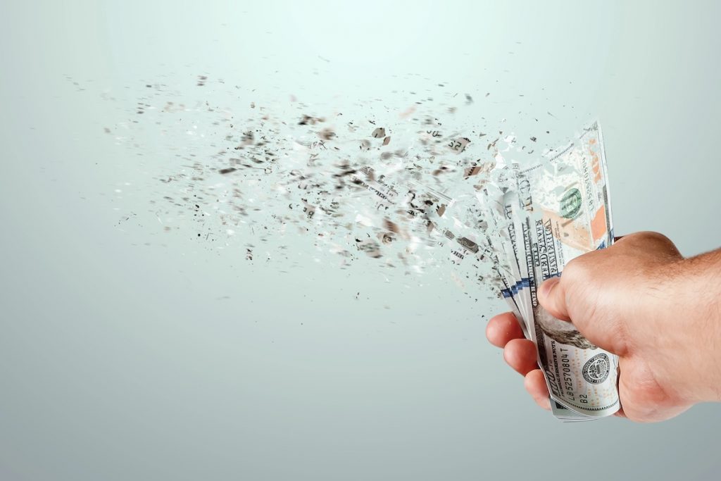 Federal Student Loan Losses Expected To Drop to 4% in 2026 Close-up of a hand holding a stack of U.S. hundred-dollar bills that are disintegrating into dust and fragments on the left side. This striking visual metaphor illustrates the concept of money losing value or disappearing, reflecting the article's discussion on federal student loan subsidies and the government's projected losses per dollar lent under new reconciliation reforms. Source: The College Investor