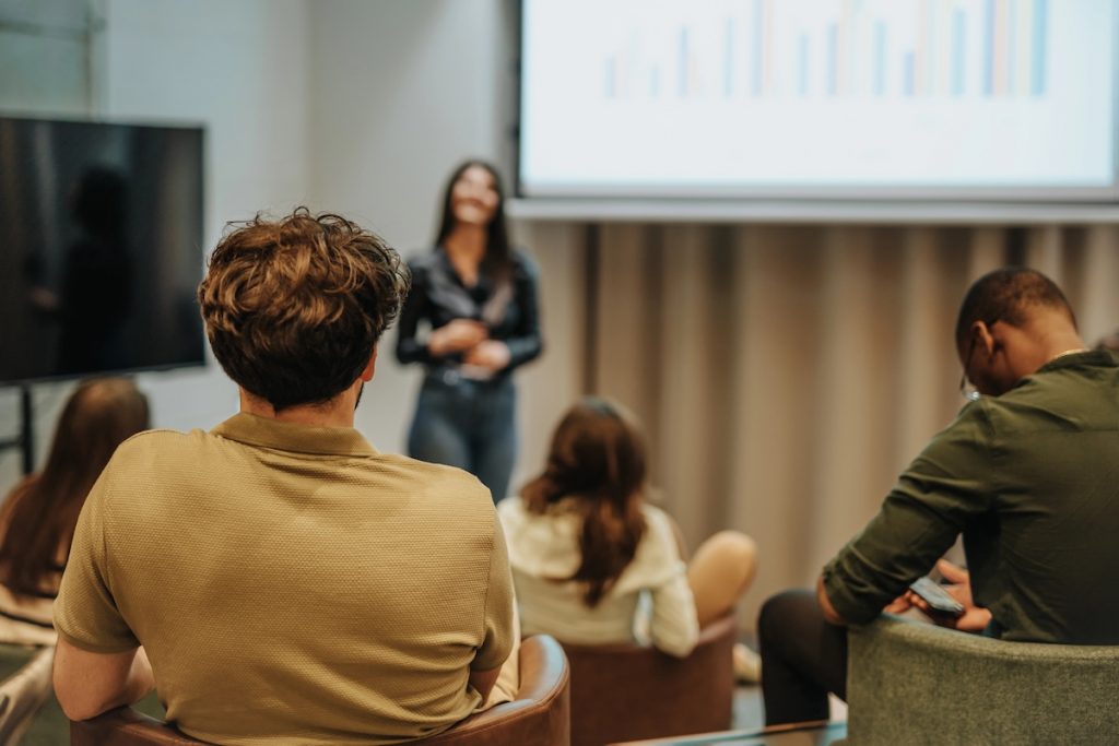 Rear view of a diverse group of adults sitting in a modern classroom, looking at a large screen displaying a colorful bar chart. A woman stands presenting, while attendees in casual attire focus on the data. This image illustrates the