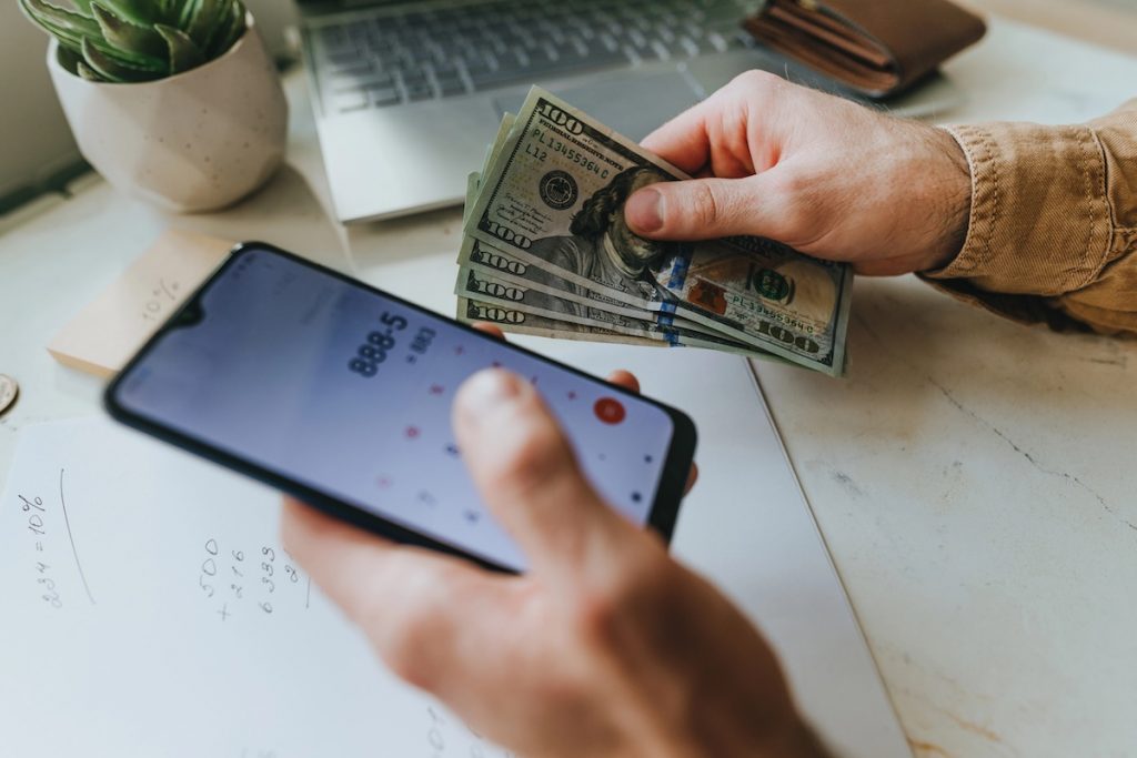 PSLF Buyback and REPAYE: How New Settlement Changes Costs Close-up of a person's hands holding a smartphone displaying a calculator app in one hand and a stack of U.S. hundred-dollar bills in the other, set over a piece of paper with handwritten math. This image illustrates the financial calculation borrowers must make regarding the PSLF Buyback program, specifically highlighting how the recent court ruling blocking the REPAYE plan could result in higher out-of-pocket costs for those seeking student loan forgiveness. Source: The College Investor