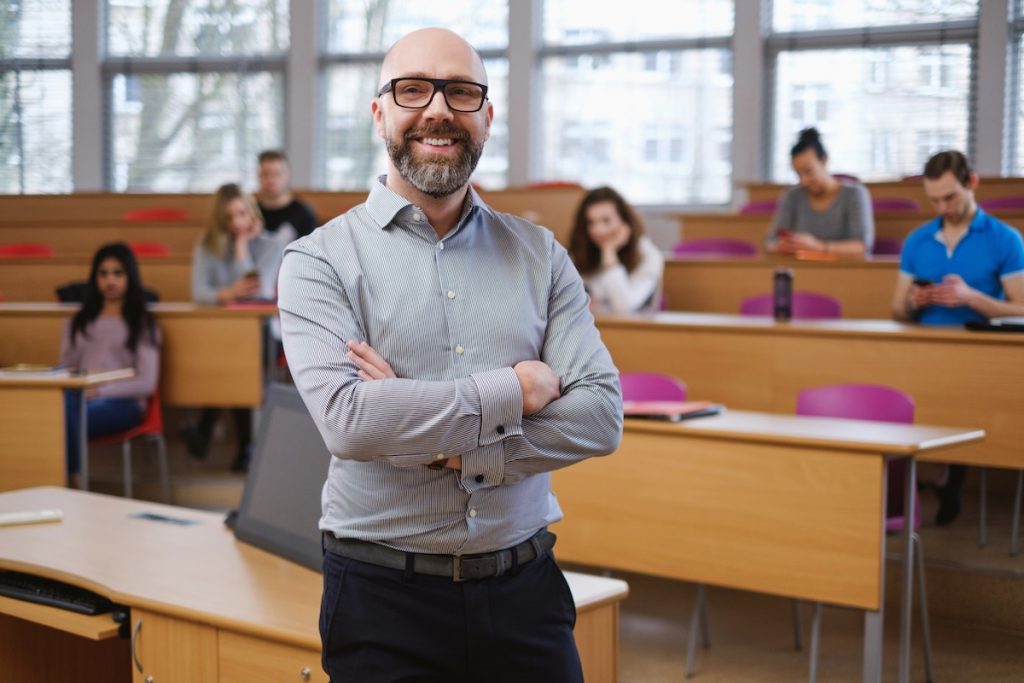 PSLF Strategy in 2026: New Employer Rule, RAP Plan, and Parent PLUS Changes A smiling male college professor with a beard and glasses stands confidently with crossed arms in the foreground of a modern university lecture hall. Behind him, several students sit at wooden desks. This image represents the public service professionals—such as educators, non-profit workers, and government employees—who must navigate complex, changing federal repayment rules outlined in the PSLF Strategy Guide to successfully achieve Public Service Loan Forgiveness. Source: The College Investor