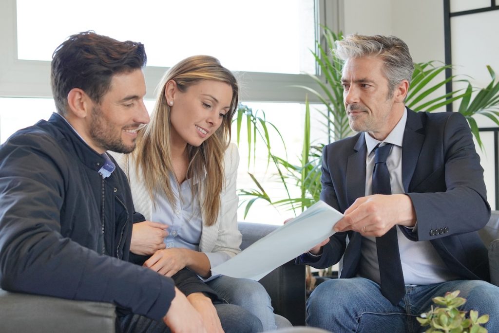 A smiling young couple sitting closely together on a grey sofa reviews financial documents handed to them by a professional advisor in a navy suit and blue tie. The three individuals are engaged in a positive discussion in a brightly lit, modern office setting with large windows and green plants. This image visualizes the consulting process for long-term financial planning, such as discussing early retirement strategies and setting up a Roth conversion ladder to access retirement funds without penalties. Source: The College Investor