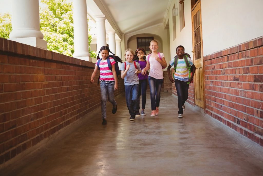 Smiling elementary school students with backpacks running down an outdoor school corridor lined with brick walls and white columns. This image illustrates the expanded utility of 529 plans, which can now be used for a broader range of K-12 educational expenses, including tuition, tutoring, and materials, with annual caps rising to $20,000 in 2026. Source: The College Investor
