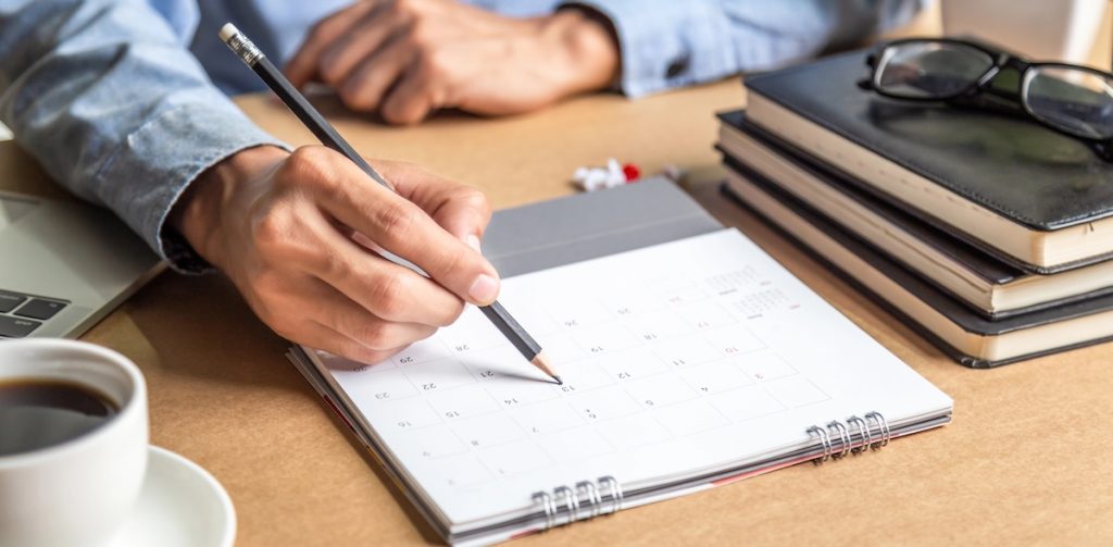 Overhead shot of a person’s hands holding a pencil, pointing to a specific date on a paper calendar laid flat on a wooden desk. Beside the calendar are a coffee cup, laptop, and a stack of books with glasses on top. This image illustrates the crucial planning needed for on time action. Source: The College Investor