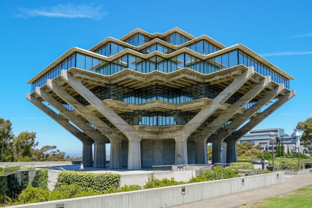 UC Admissions Board Scraps SAT Review Plan One Day Before Regents Meet The iconic Geisel Library at the University of California San Diego stands majestically against a bright blue sky, its unique brutalist architecture resembling hands holding up a stack of books. This striking eight-story concrete and glass structure, with its geometric patterns and expansive windows reflecting the surroundings, is framed by lush green landscaping and pathways, hinting at the vibrant campus environment. The image visually anchors the article discussing concerns about UCSD freshmen's declining math skills and the university's response with remedial courses, raising questions about admissions practices, test-optional policies, and grade inflation amidst record demand for college admissions.