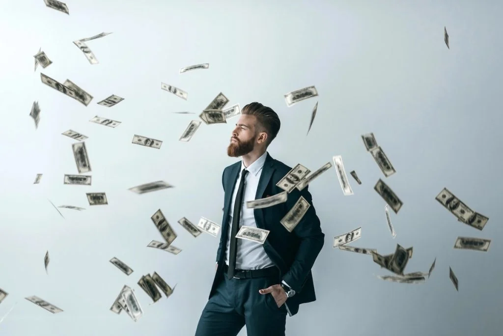 Bearded man in a dark business suit standing confidently with his hand in his pocket while US dollar bills rain down around him against a gray background. This illustrates the potential for wealth accumulation and passive income through high-yield savings accounts, which are currently offering interest rates up to 5.00% APY as of November 2025, allowing savers to maximize returns despite fluctuating market conditions. Source: The College Investor