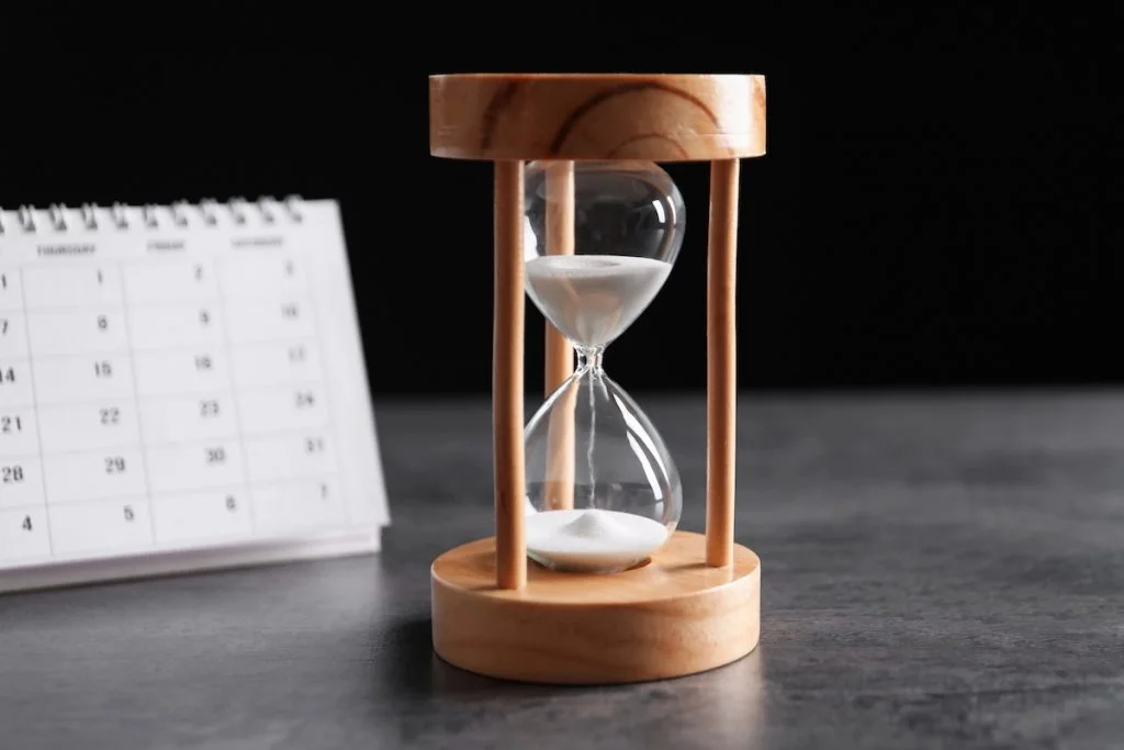 Close-up of a wooden hourglass with sand running out, sitting next to a white paper calendar on a dark surface. This image visualizes the urgency of the upcoming July 1, 2026, deadline for federal student loan borrowers—especially Parent PLUS holders—to consolidate their loans, or risk permanently losing access to income-driven repayment plans and forgiveness programs. Source: The College Investor