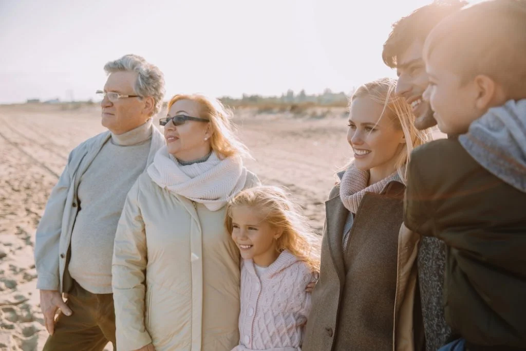 Parents Spend $1,474/Month on Adult Kids vs. $673 on Retirement A multi-generational family of six, including grandparents, parents, and two young children, stands together on a sunny beach, smiling and looking out toward the horizon. They are dressed in warm, neutral-toned fall or winter coats and scarves. This image visually represents the concept of family support and interconnectedness, reflecting the article's focus on how rising student debt and housing costs are increasingly causing young adults to rely on their parents for financial help, impacting multiple generations' financial futures. Source: The College Investor